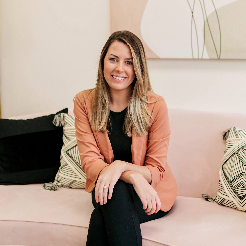 Blonde woman in a peach blazer sitting on a light pink sofa and smiling.
